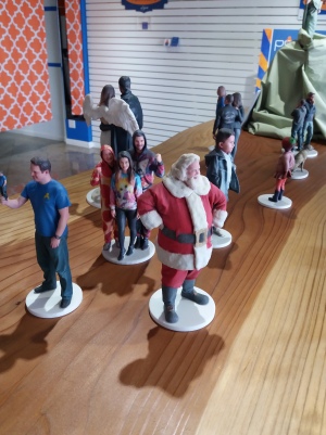 A plain wooden shelf features a selection of a dozen figurines, including children, families in matching outfits, and Santa Claus.