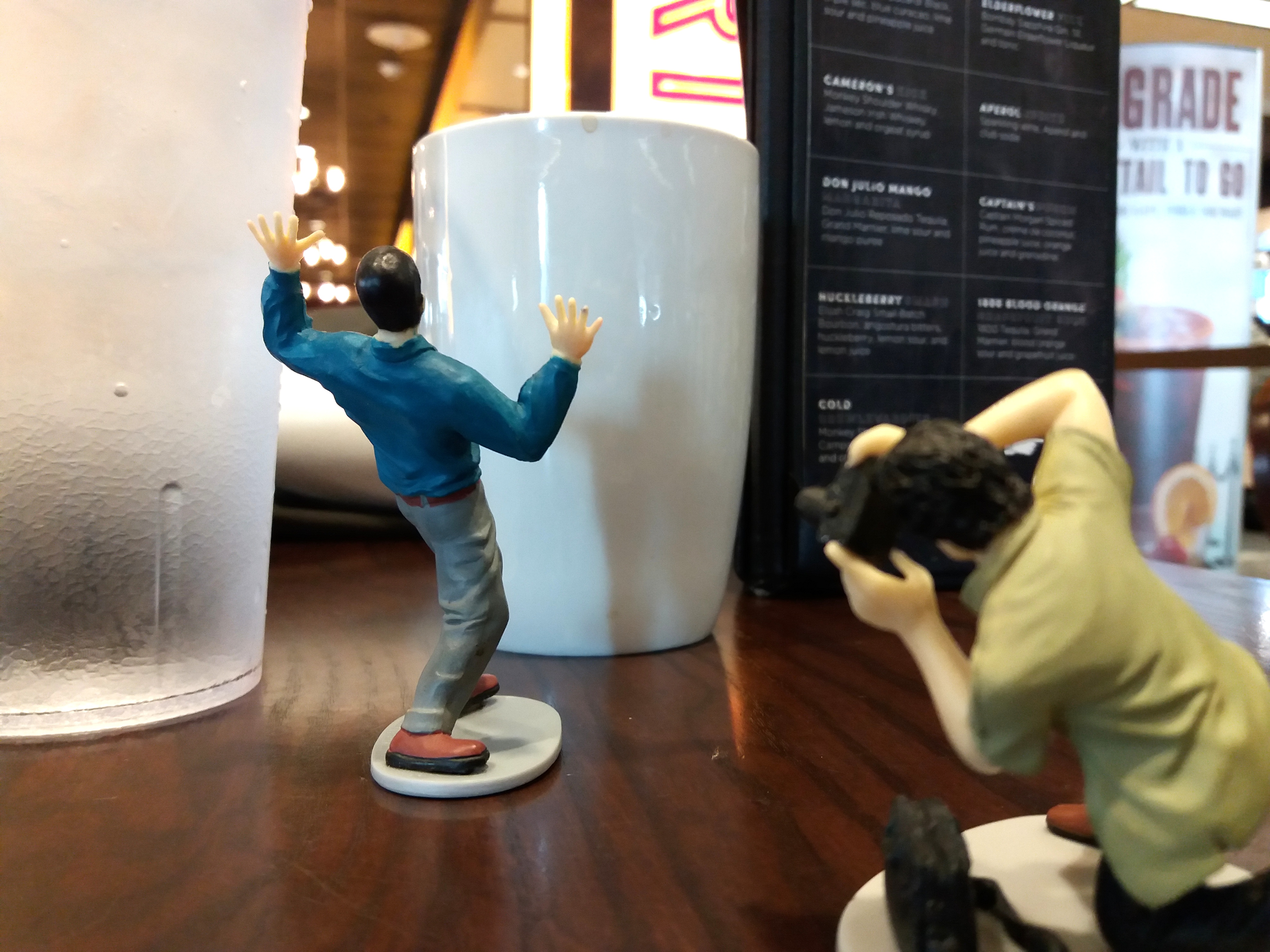 A tiny man rears in horror at a normal-sized coffee mug; behind him, a tiny paparazzi takes a photo of the spectacle. This is all staged on a table at a restaurant, with a standing menu in the background.