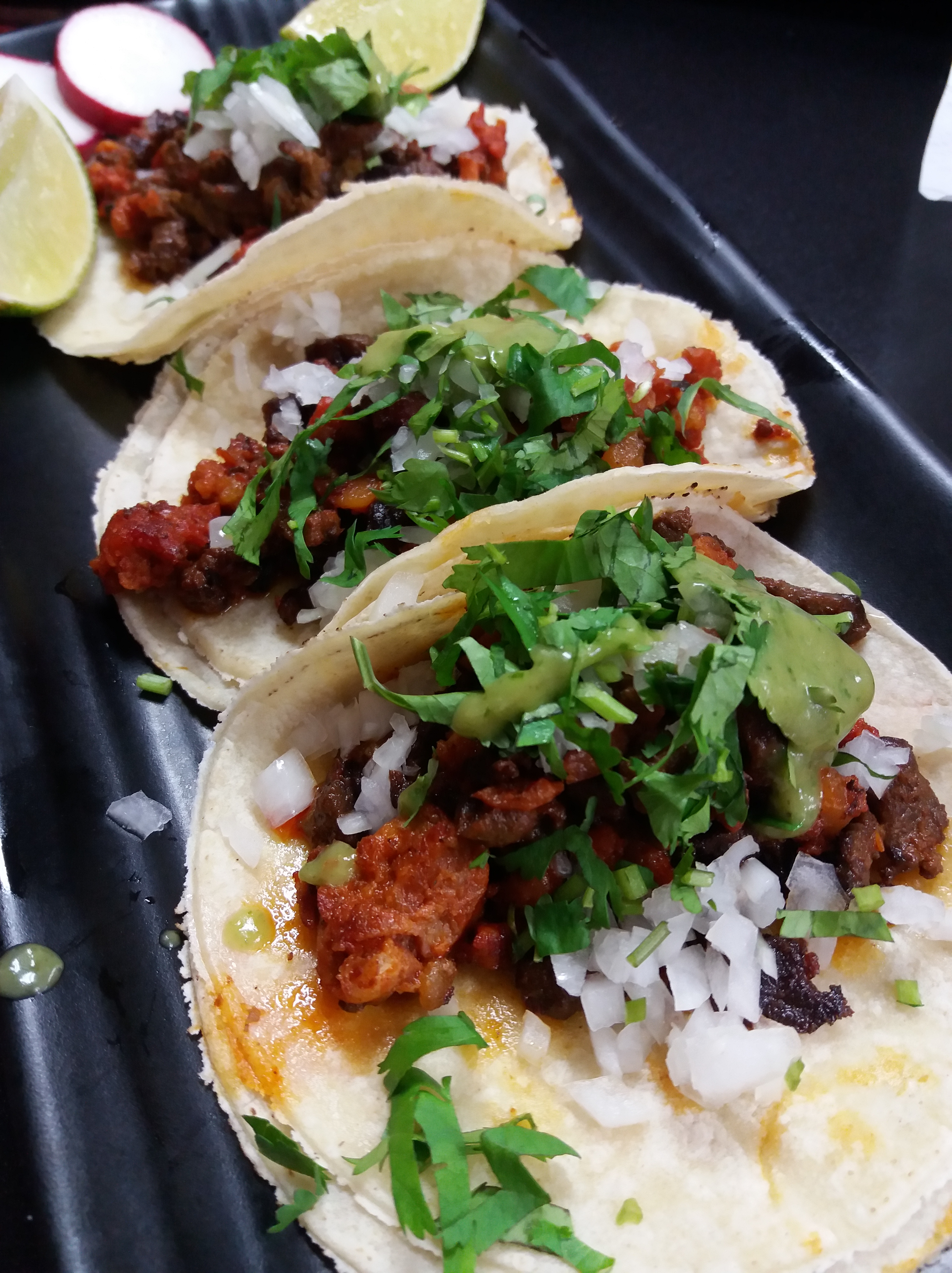 A set of three soft-shell tacos on a black stoneware tray. They all feature fresh minced onions, various grilled meats with sauces, verde salsa and lots of cilantro.