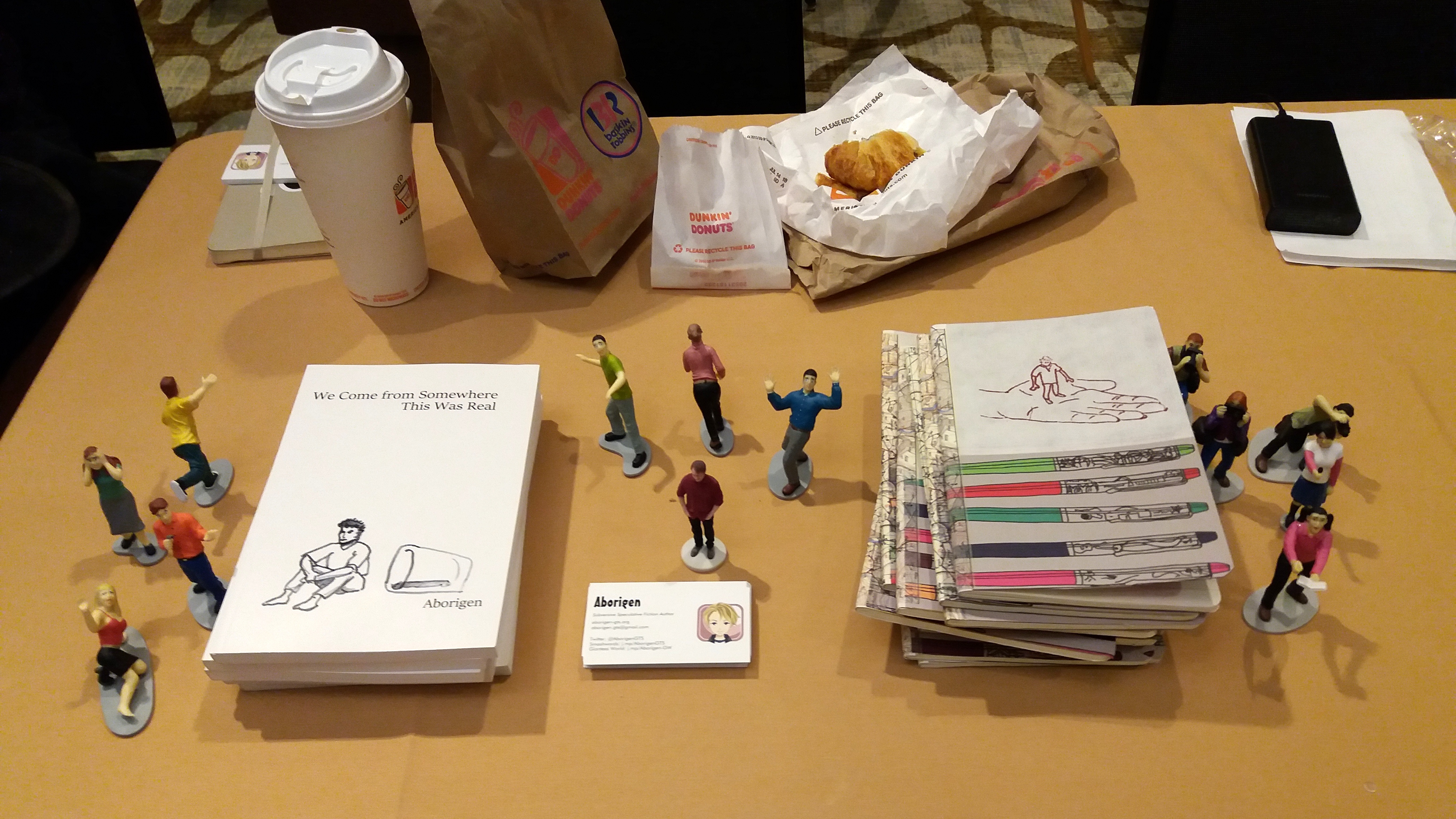 On a beige tablecloth, Aborigen's books of short stories are stacked on the left and his custom notebooks on the right. Around these stand tiny, frightened figurines. Behind it all is his breakfast from Dunkin Donuts.