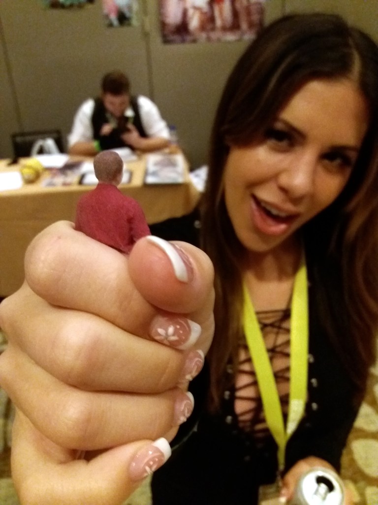 Giantess Katelyn, attending SizeCon, clutches a 3" tall Aborigen statuette in her manicured fist and leers at the camera.