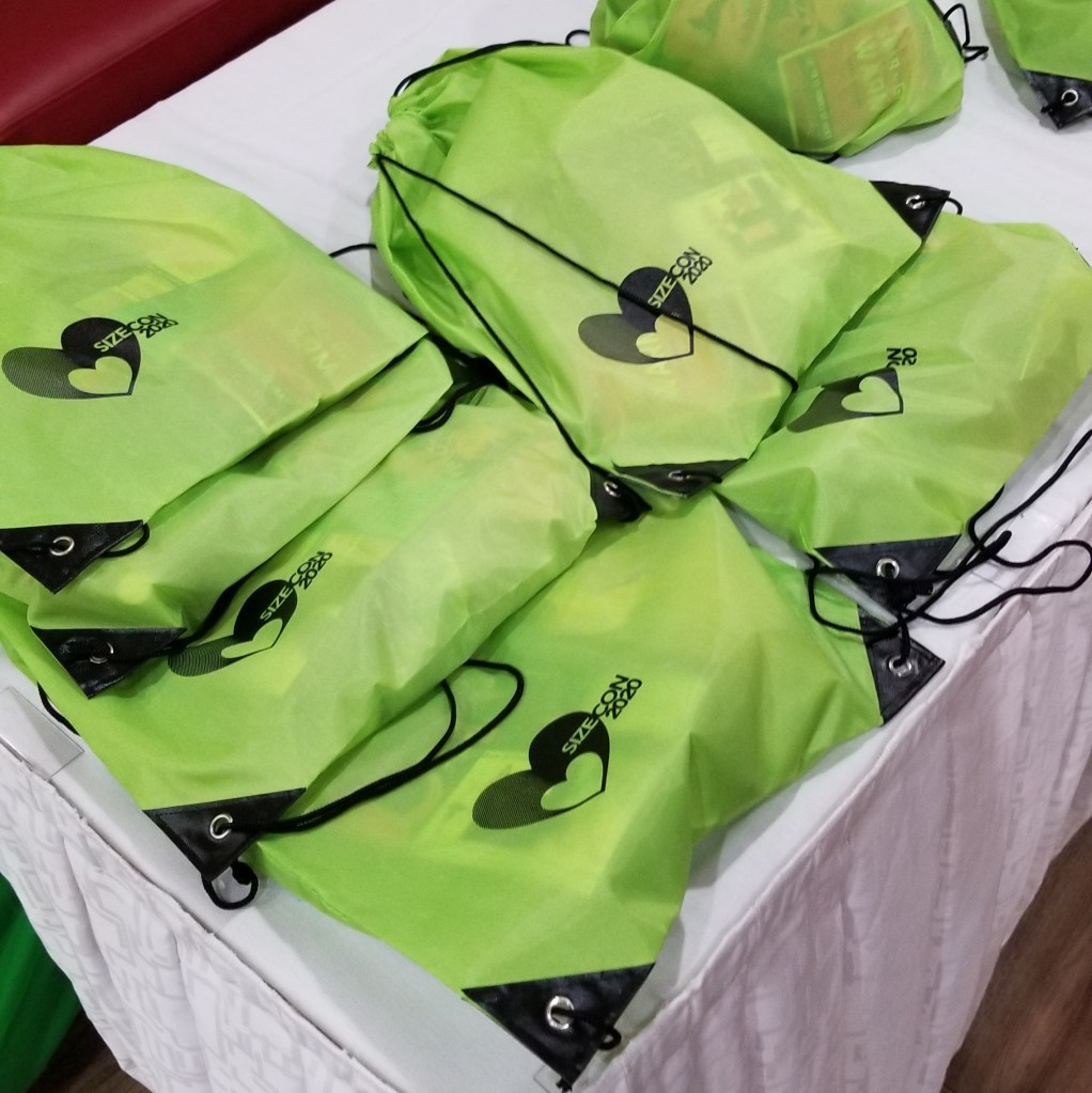 Several green nylon string backpacks marked "SizeCon" rest on a white tablecloth.