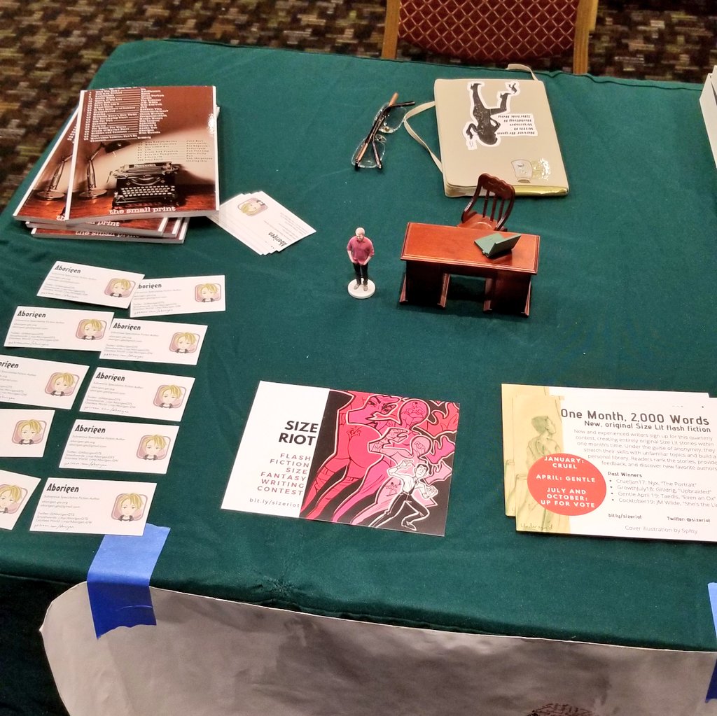 Aborigen's cluttered booth at SizeCon: business cards, Size Riot postcards, copies of Small Print, and a miniature desk and chair.