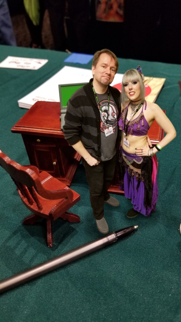 Tiny Aborigen and Morgana stand in front of his tiny desk on a gigantic table, a pen lying at their feet.