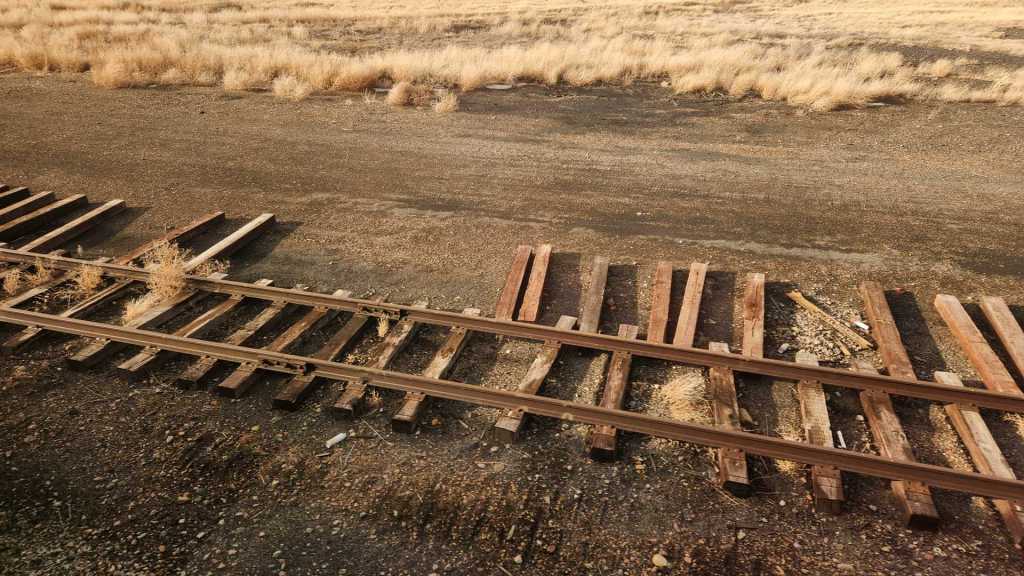 A set of surplus train tracks with railroad ties lying in disorder, with brush in the background.
