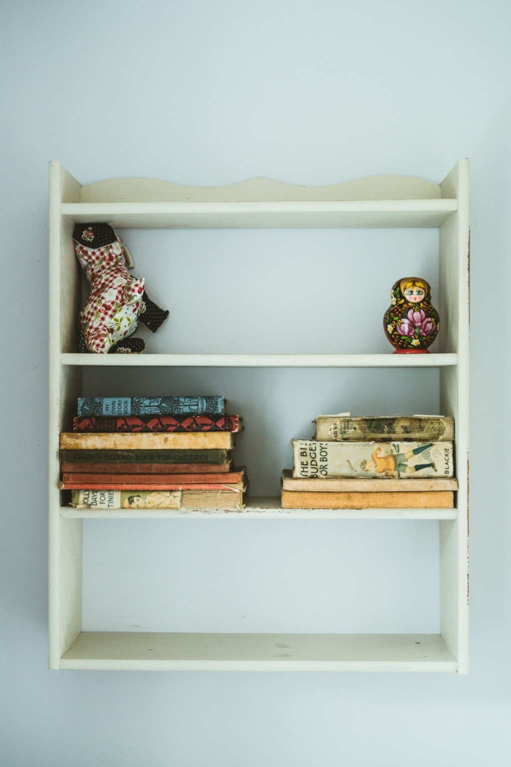 A small white bookshelf hung from a white wall. The top shelf has two small dolls, the second has two piles of vintage children's books, and the third is bare.