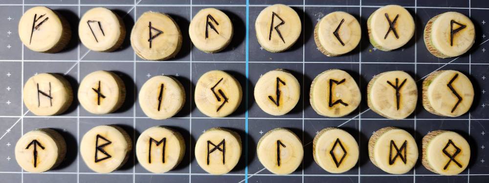 Three rows of eight wooden disks bearing Elder Futhark runes, resting on a Cricut mat.