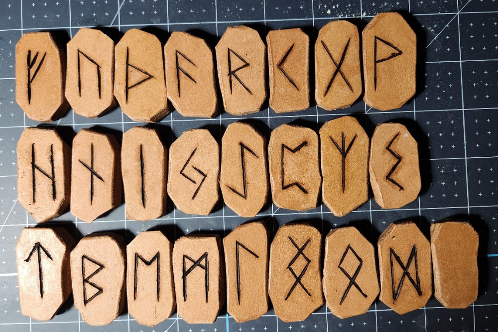 Three rows of leather tiles bearing Elder Futhark runes, resting on a Cricut mat.