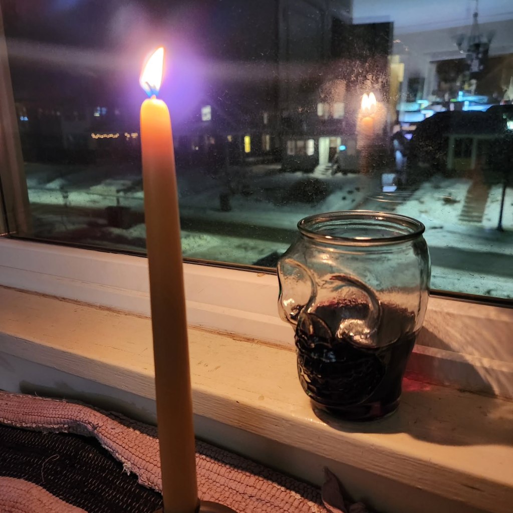 A tall beeswax candle next to a skull-shaped glass of wine on a windowsill, overlooking a wintry neighborhood.